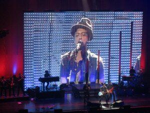 Bruno Mars performing live with his likeness projected on a big screen behind the band.